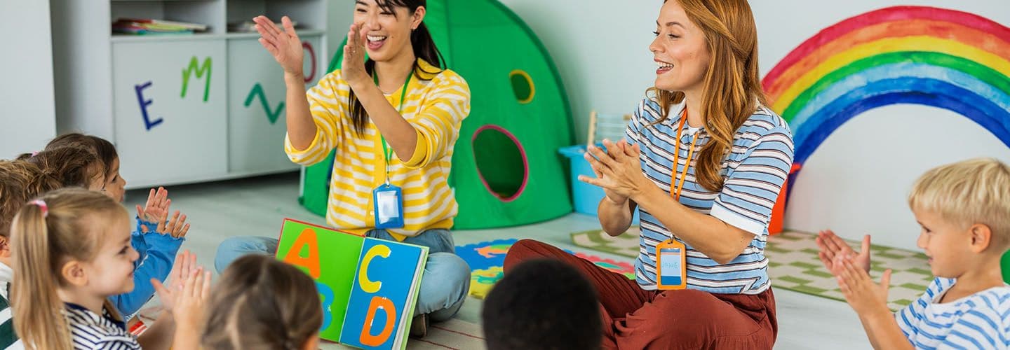 two daycare teachers sit on the floor with their students while reading them a story